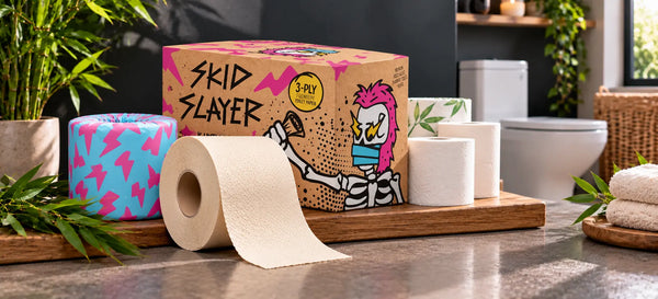 Skid Slayer cream bamboo toilet paper roll, cyan-pink wrapper and kraft box beside other toilet paper rolls on a wooden bathroom display.