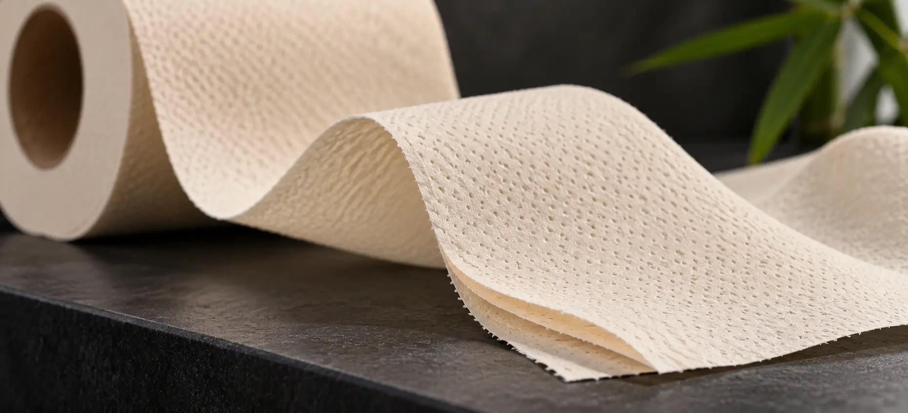 Close-up of cream bamboo toilet tissue with soft folds, fine embossed texture and separated layers on a dark counter.