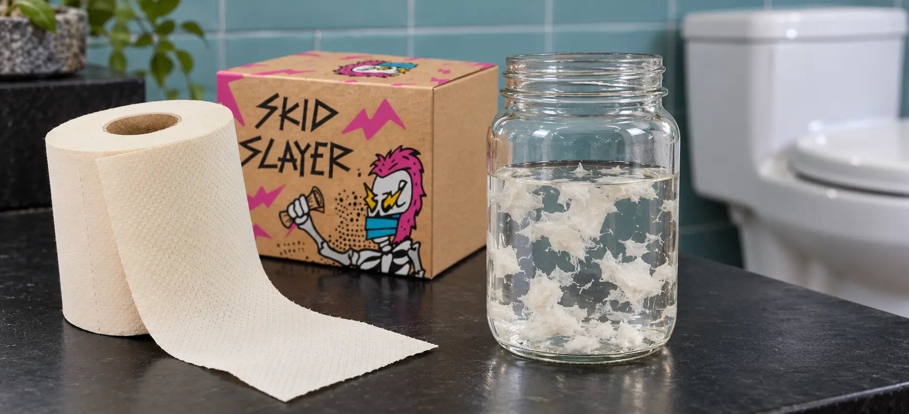 Cream bamboo toilet paper roll beside a Skid Slayer carton and a glass jar containing tissue fragments in water.