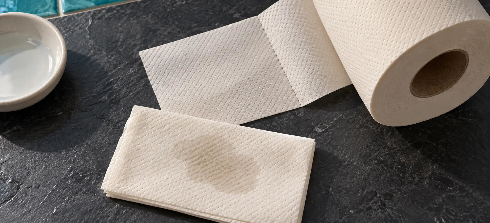 Skid Slayer bamboo toilet paper beside folded tissue with a damp patch on a dark bathroom counter.
