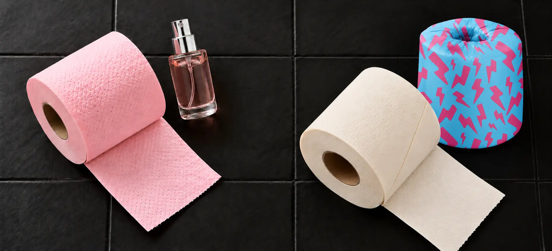 Pink toilet paper beside a perfume bottle, with a cream bamboo roll and cyan-pink wrapped Skid Slayer roll on black tiles.